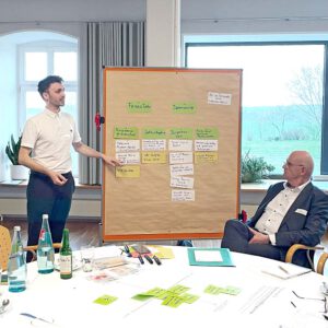 Eine Person steht an einer Moderationspinnwand mit Moderationskarten und zeigt darauf. Davor sitzt eine Person und schaut zu. Im Bildvordergrund steht ein Tisch mit Workshop- und Moderationsmaterialien
